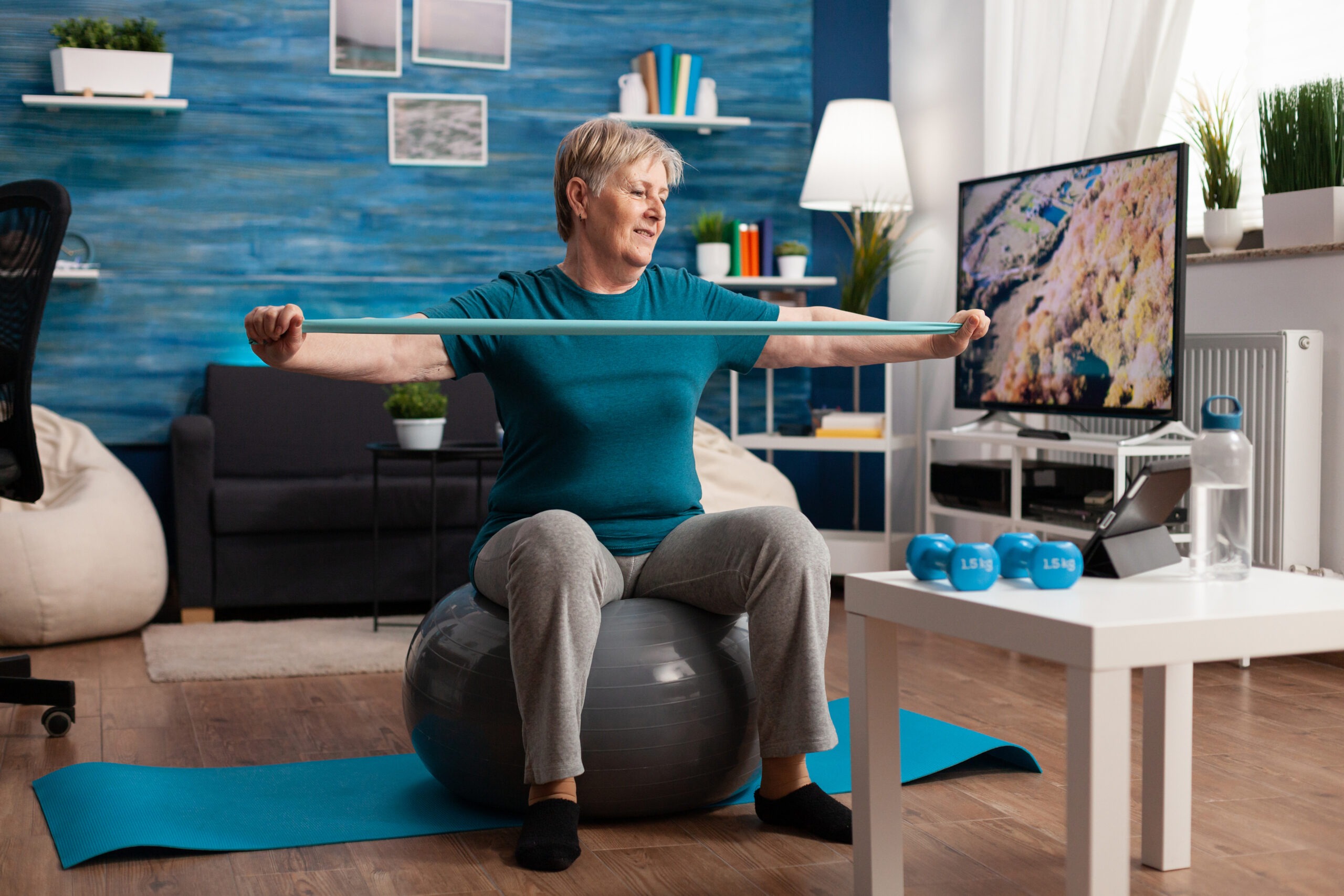 Retired senior female sitting on fitness swiss ball with exercise band in living room doing online virtual fitness workout streching arm muscles using aerobics elastic band.